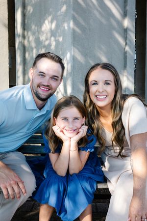 A smiling family of three poses on steps, with a young girl in blue flanked by her parents in casual attire.