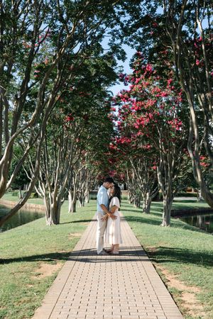 Couple embracing on a brick pathway lined with blooming pink crepe myrtle trees in a lush green park.