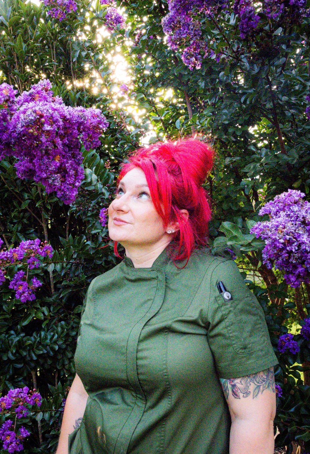 Woman with red hair in green chef coat stands among purple flowering bushes, looking up serenely.