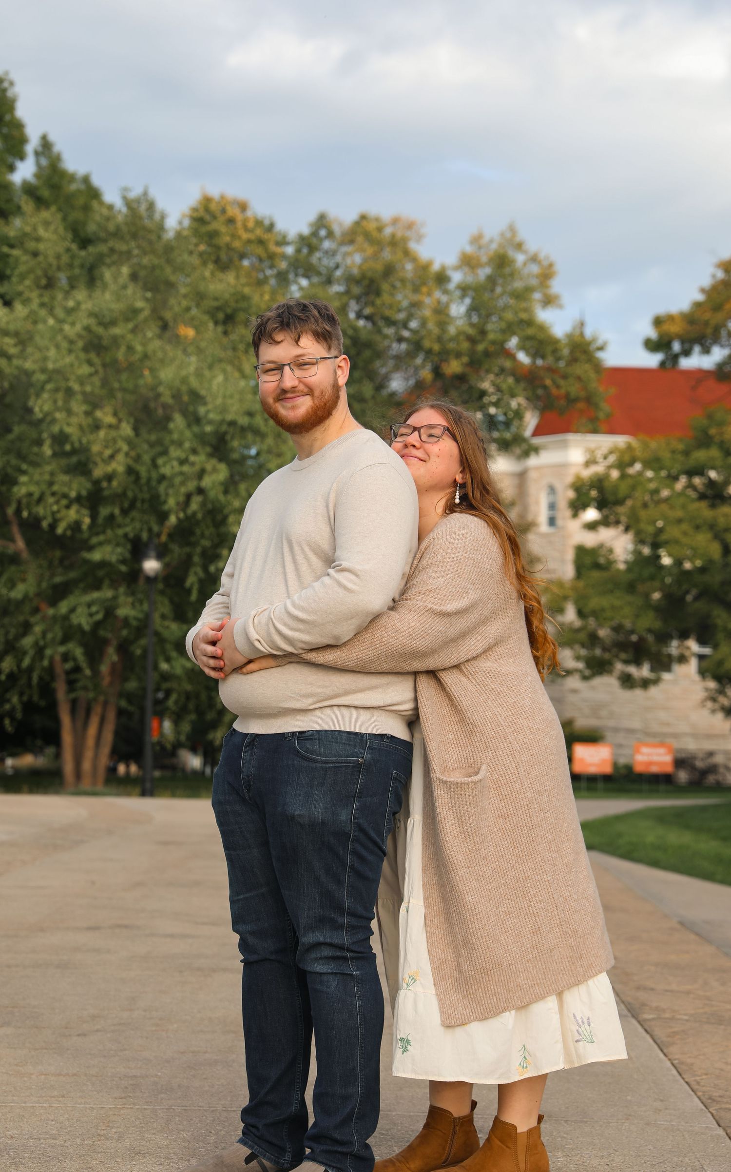 A couple poses outdoors, woman hugging man from behind, both wearing glasses and casual fall attire near a university building.
