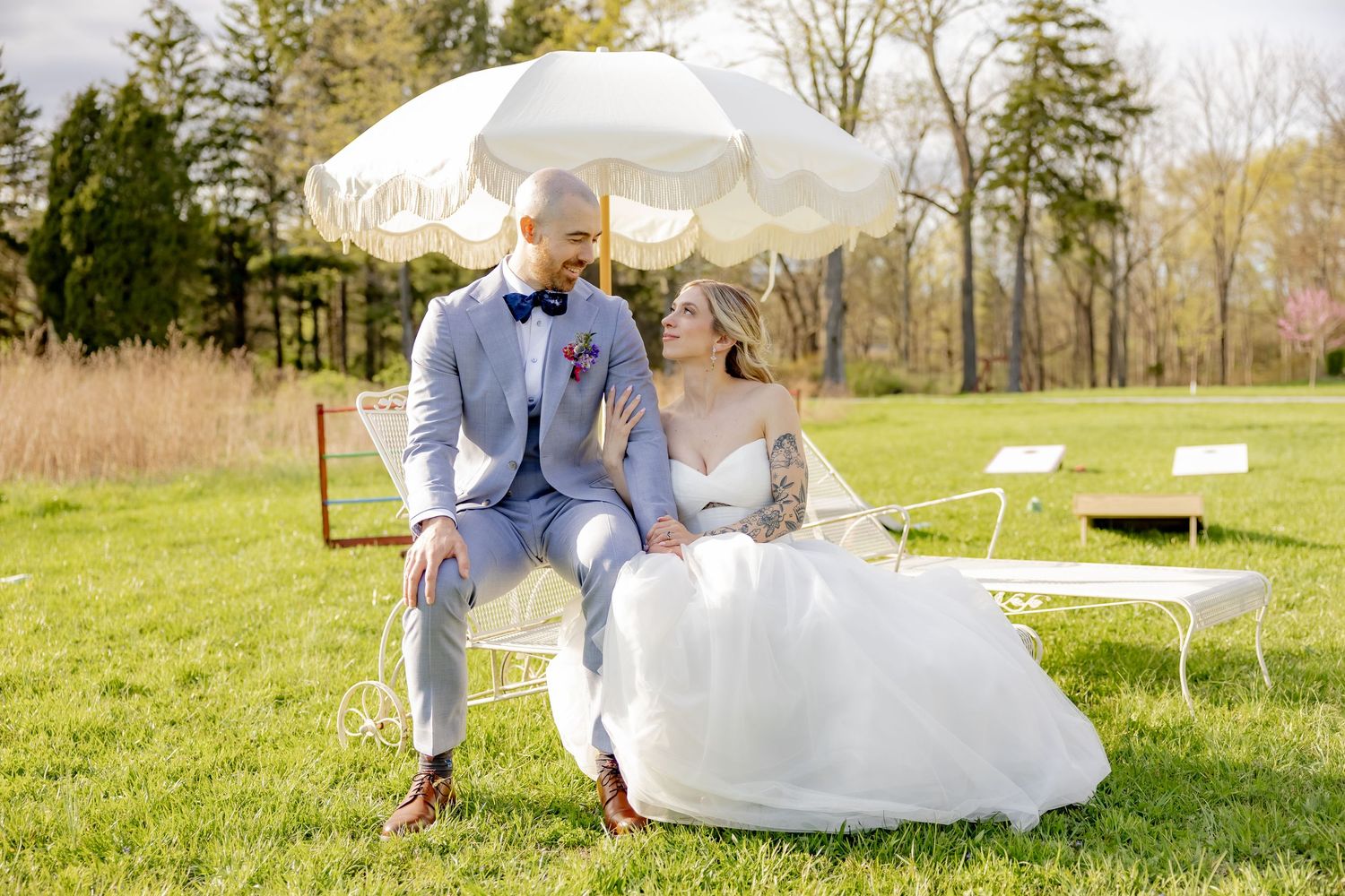 Bride and groom sitting together on white garden furniture under a decorative umbrella on a lush green lawn.