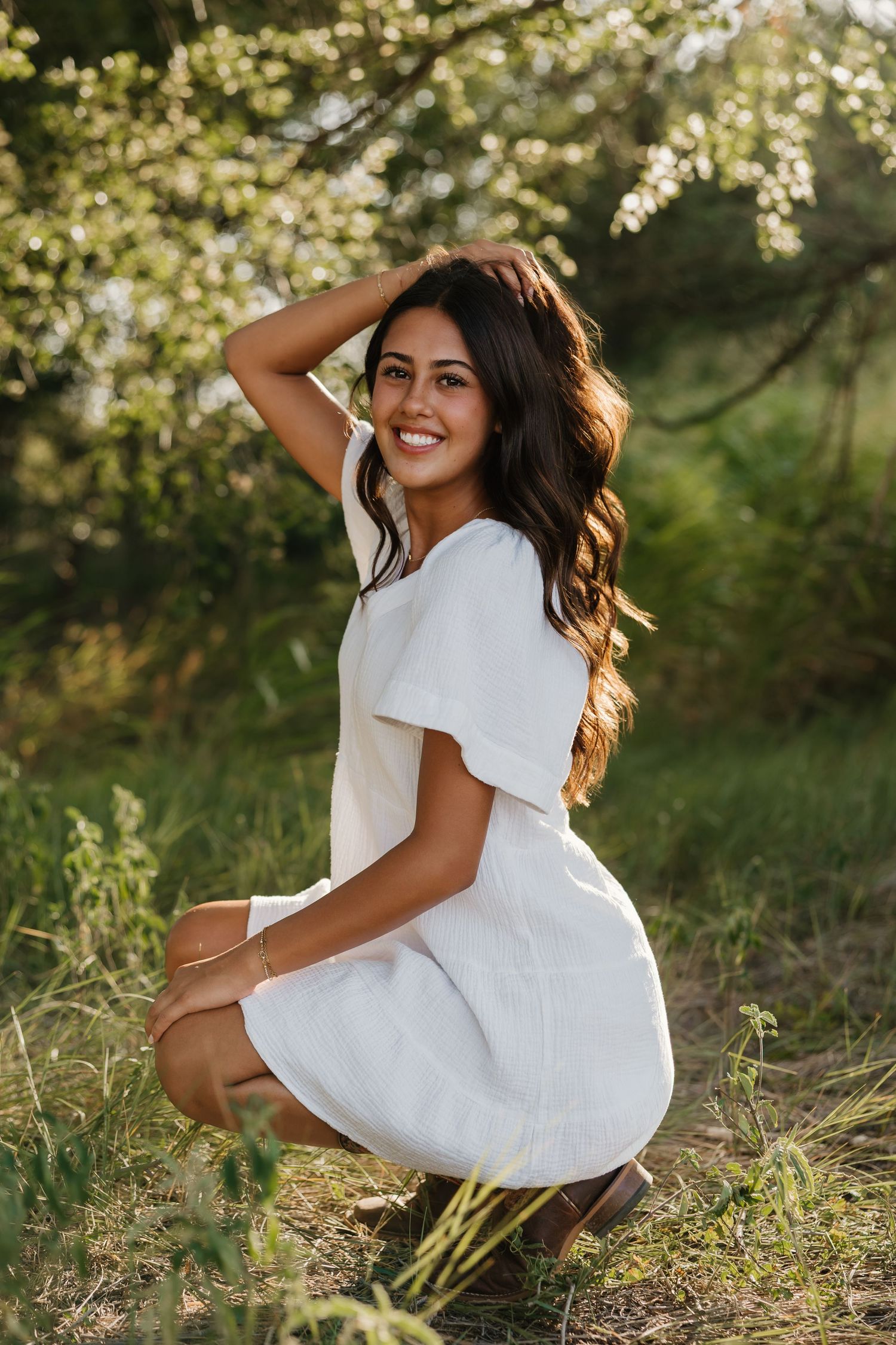 A girl smiling and playing with her hair in a bright and green field posing for senior photos in white dress and boots