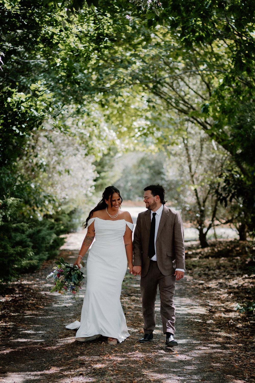 A couple in wedding attire walks together on a wooded path surrounded by green trees and dappled sunlight.