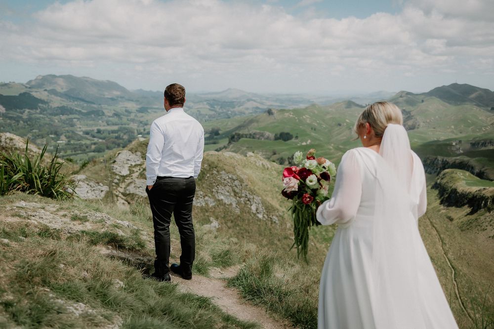 Couple in wedding attire walking along scenic mountain path with rolling hills and cloudy sky in background.