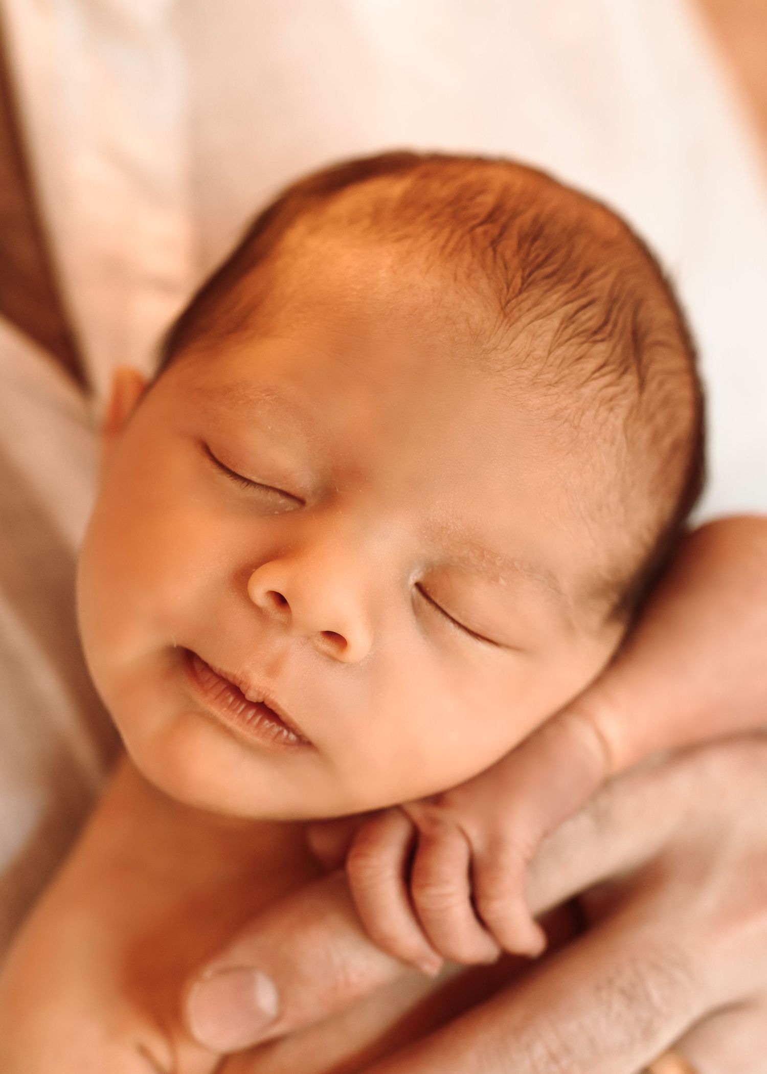 A peaceful sleeping newborn baby with wispy dark hair rests against soft white fabric.