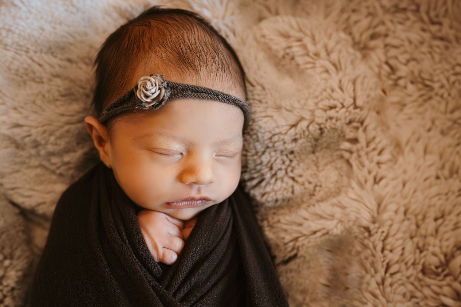 Newborn baby sleeping peacefully on soft beige fur blanket with delicate headband accessory at newborn session