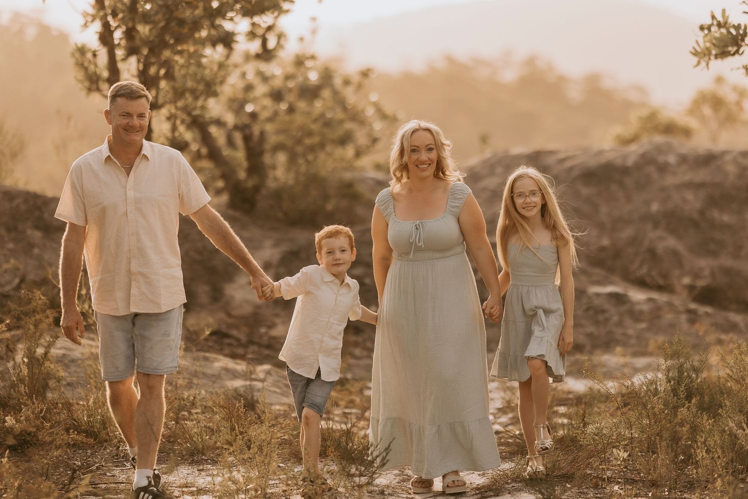 Family of four walking through a sunny wildflower field, parents holding toddler hands, golden hour light.