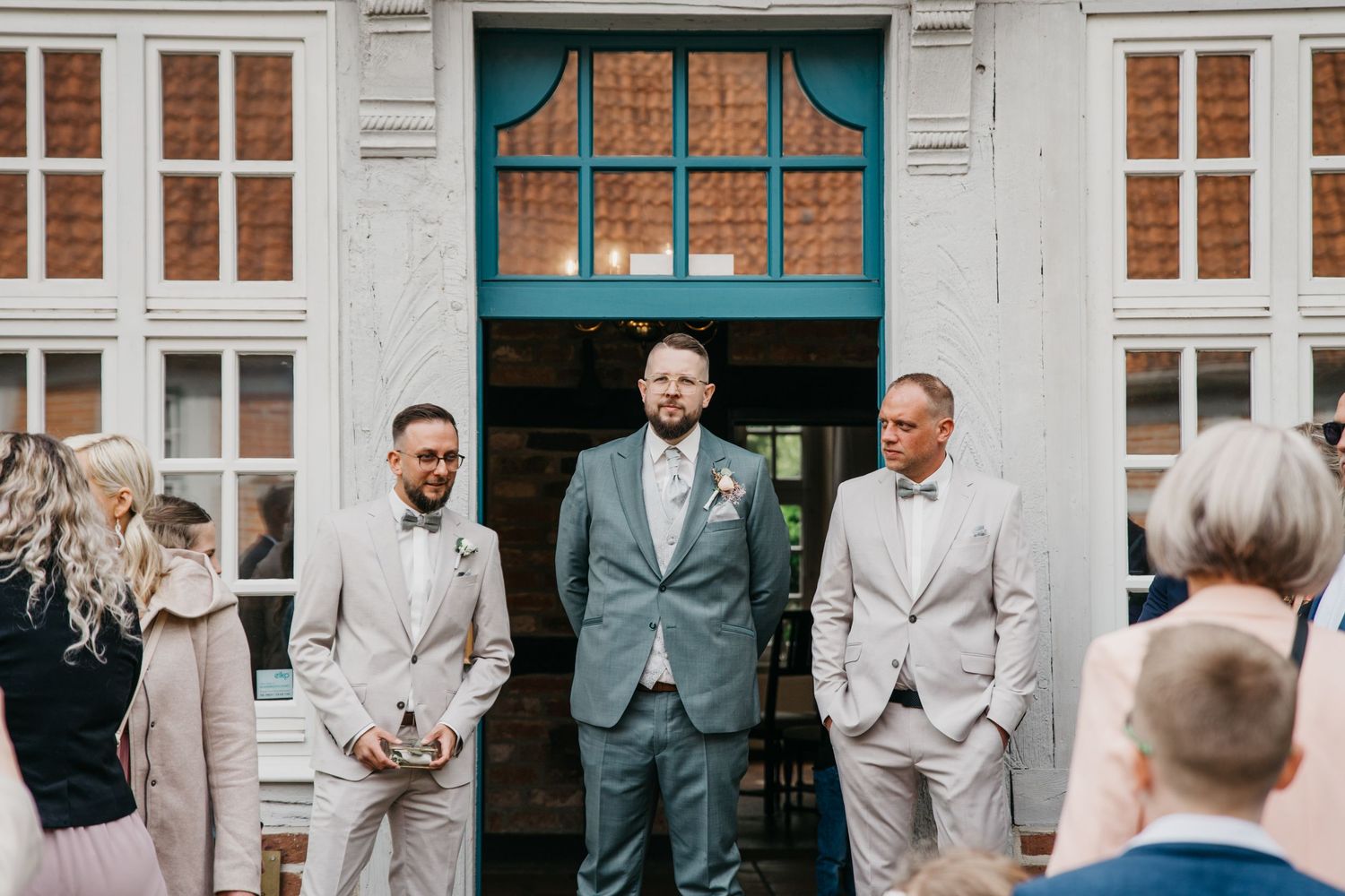 Groom in a gray suit stands in a doorway flanked by two groomsmen as wedding guests gather in the foreground.