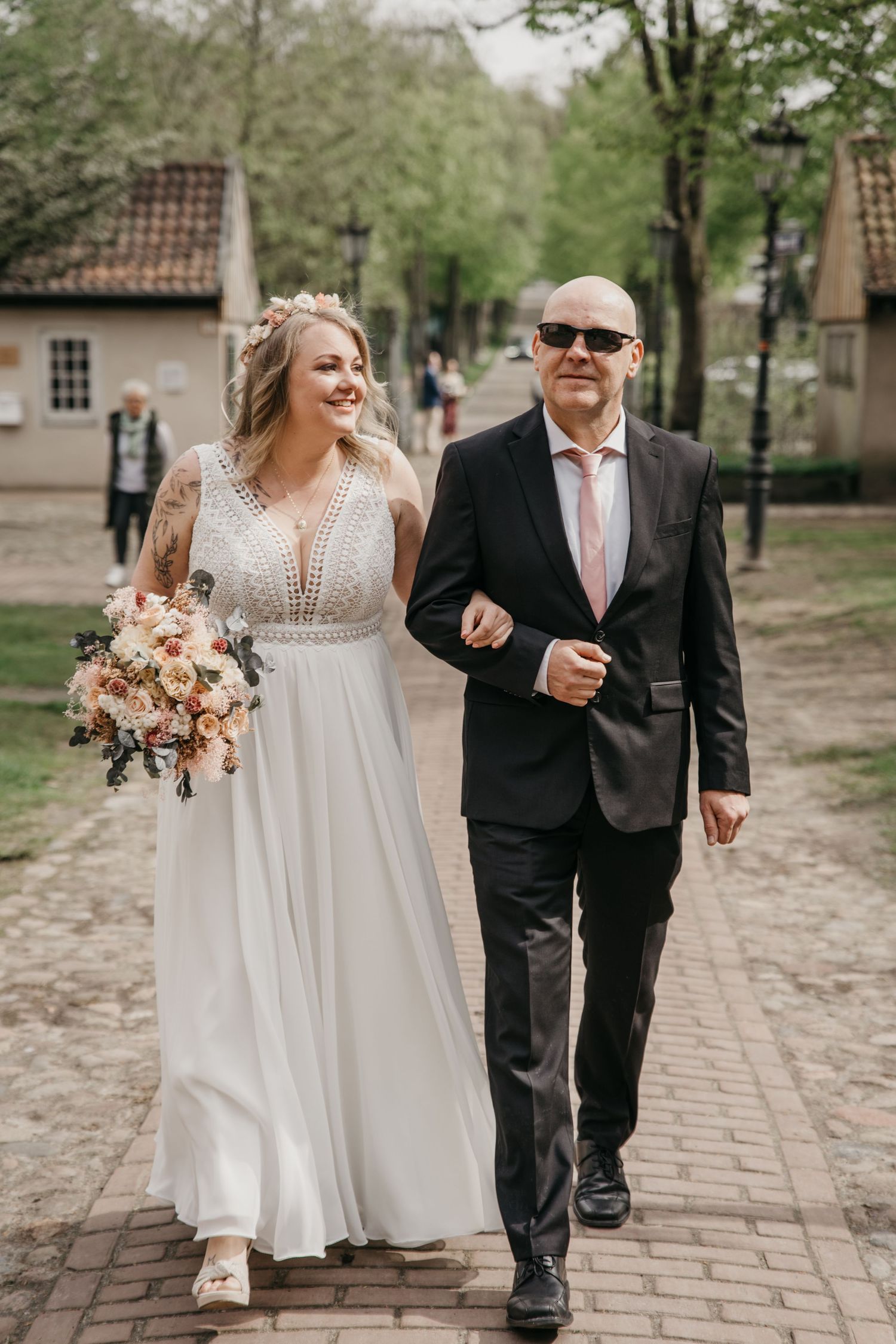 Smiling bride in a white lace dress holding a bouquet walks arm in arm with a man in sunglasses along a brick path.