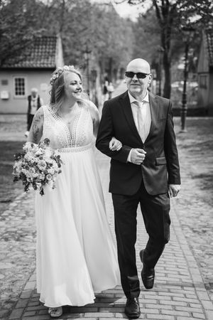Black and white photo of a smiling bride with a floral crown and bouquet walking arm in arm with a man in sunglasses.