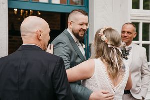 A bald man in a dark suit embraces a bride in a lace wedding dress with a floral hair crown as the groom and a guest look on.