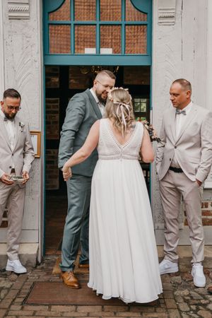 A bride in a white lace gown holds hands with the groom in a green suit at a teal doorway, flanked by two groomsmen in beige suits.