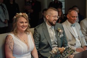 A joyful bride in a lace dress with a flower crown sits beside her bearded groom in a green suit, holding a dried flower bouquet.