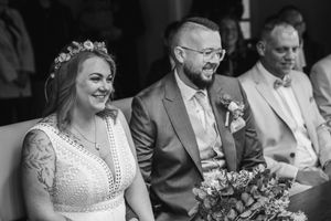 A smiling bride with a floral crown and a bearded groom in glasses sit side by side holding a bouquet during their wedding ceremony.