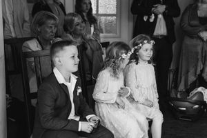 A black and white photo of a boy and two flower girls in floral crowns sitting attentively during a wedding ceremony.