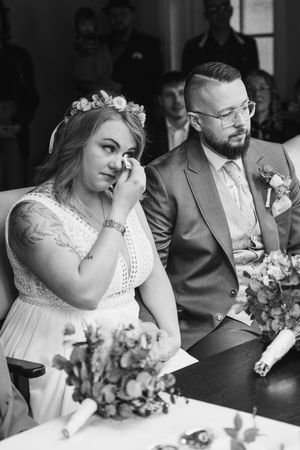A black and white photo of an emotional bride wiping a tear while seated beside her groom at the wedding ceremony table.
