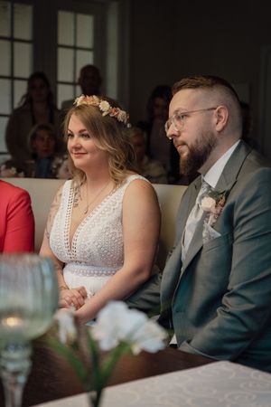 A bride with a floral hair crown and a bearded groom in glasses and green suit watch the wedding ceremony with a vase in the foreground.
