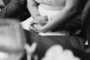 A black and white close-up of the bride and groom holding hands during their wedding ceremony, with the bride's tattooed arm visible.