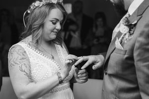 Black and white photo of a bride in a lace dress and flower crown as the groom places a wedding ring on her finger.