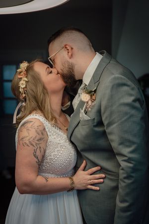 A bride with a floral crown and a groom in a gray suit with glasses share a romantic kiss during their wedding ceremony.