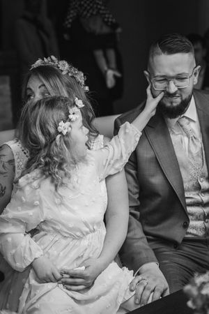 Black and white photo of a little flower girl playfully touching the groom's cheek as he smiles beside the bride at the ceremony.