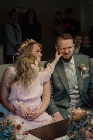 A flower girl in a floral headband playfully pats the groom's cheek while sitting on the bride's lap during the wedding ceremony.