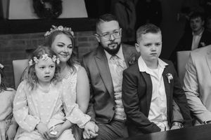 A bride, groom, young girl and boy sit together holding hands during a wedding ceremony, with guests visible in the background.