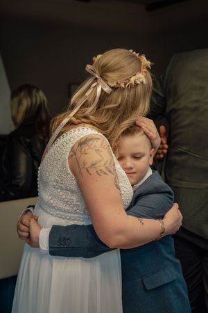 A bride with a flower crown and ribbon tenderly hugs a young boy in a blue suit during her wedding day.