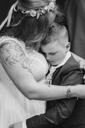 Black and white close-up of a bride with a leafy arm tattoo embracing a young boy in a suit with his eyes closed.