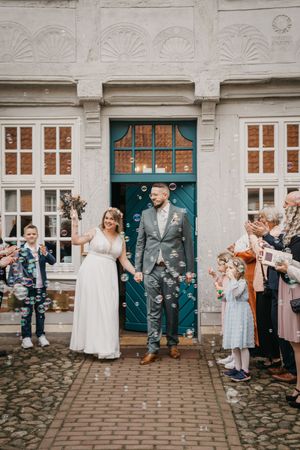 Newlyweds hold hands outside a historic building with a teal door as guests blow bubbles and cheer around them.