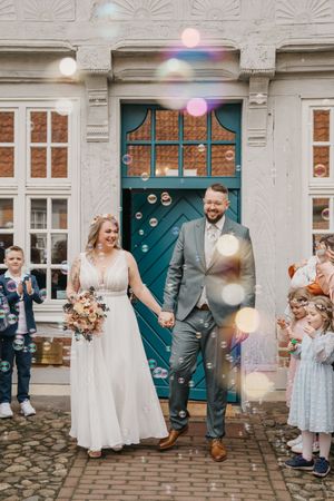 A smiling bride with a bouquet and groom in a gray suit walk hand in hand through floating bubbles outside a teal door.