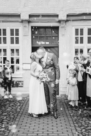 Black and white photo of newlyweds kissing on a brick path surrounded by bubbles while wedding guests watch and celebrate.
