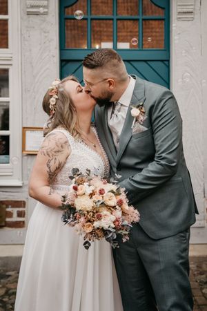Bride in a white lace gown and groom in a gray suit kiss in front of a teal door, holding a pastel bouquet.