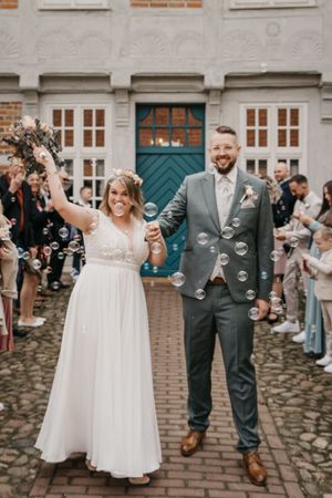 Smiling newlywed couple walks hand in hand through floating soap bubbles as wedding guests cheer on both sides.