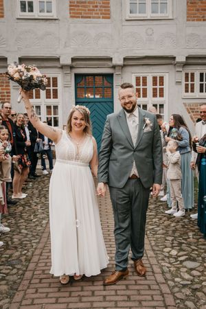 Happy bride raises her bouquet while holding hands with her bearded groom as wedding guests line the brick path.