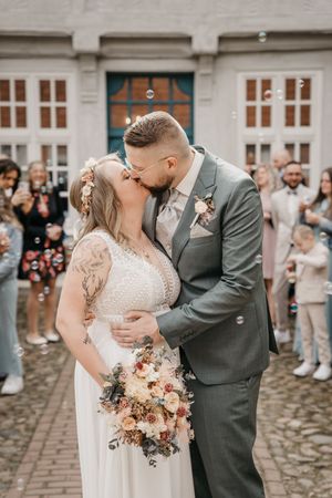 Newlywed couple kisses on a cobblestone courtyard surrounded by soap bubbles, with the bride holding a dried flower bouquet.