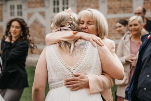 Bride in a beaded lace wedding dress is warmly embraced by a smiling older woman while guests look on.