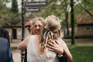 Bride with a floral hair crown and ribbon hugs a smiling woman wearing glasses at an outdoor wedding celebration.