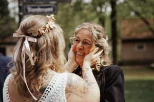 Emotional older woman in glasses smiles as the tattooed bride in a lace dress gently holds her face.