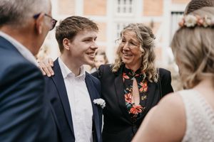 Smiling young man in a navy suit stands beside a joyful woman in a floral blouse as wedding guests mingle.