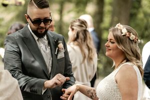Groom in sunglasses and a gray suit holds hands with the bride wearing a flower crown during an outdoor wedding ceremony.