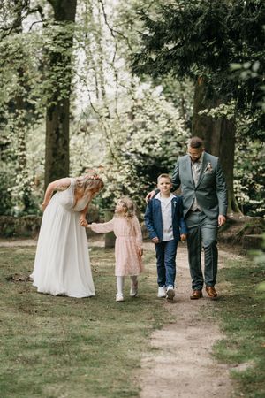 Bride in a flowing white gown and groom in a green suit walk hand in hand with two children along a sunlit forest path.