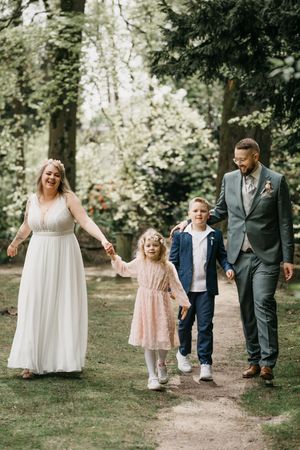 Smiling bride and groom walk a woodland trail holding hands with a little girl in a pink dress and a boy in a navy blazer.