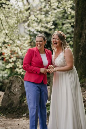 Two joyful brides laugh together, one in a red blazer and blue trousers, the other in a lace white wedding dress, in a garden.