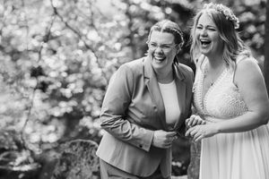 Black and white photo of two laughing brides, one in a blazer and glasses, the other in a lace wedding gown with a flower crown.