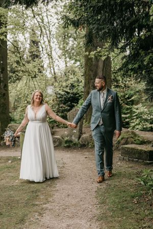 Bride in a white dress and groom in a sage green suit hold hands while walking down a gravel path through a leafy forest.