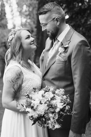 Black and white portrait of a bride with a floral hair crown gazing up at her bearded groom in a suit, holding a white bouquet.