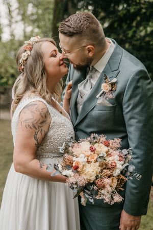 Groom in a green suit leans in to kiss his bride, who wears a flower crown and holds a pink and peach bouquet in a garden.