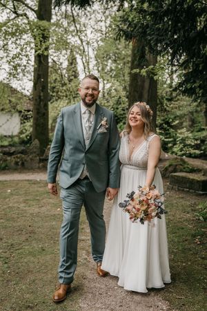 Smiling groom in a green suit and bride in a white gown holding a colorful bouquet stand together on a path in a wooded park.