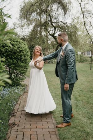 Groom in a green suit twirls his smiling bride in a white wedding dress on a brick garden path surrounded by lush greenery.
